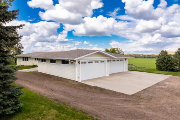 A single-story white house with a two-car garage sits on a spacious lot with a gravel driveway, green lawn, trees, and scenic views—perfect as a hunting property or cattle ranch under the open fields and blue sky.