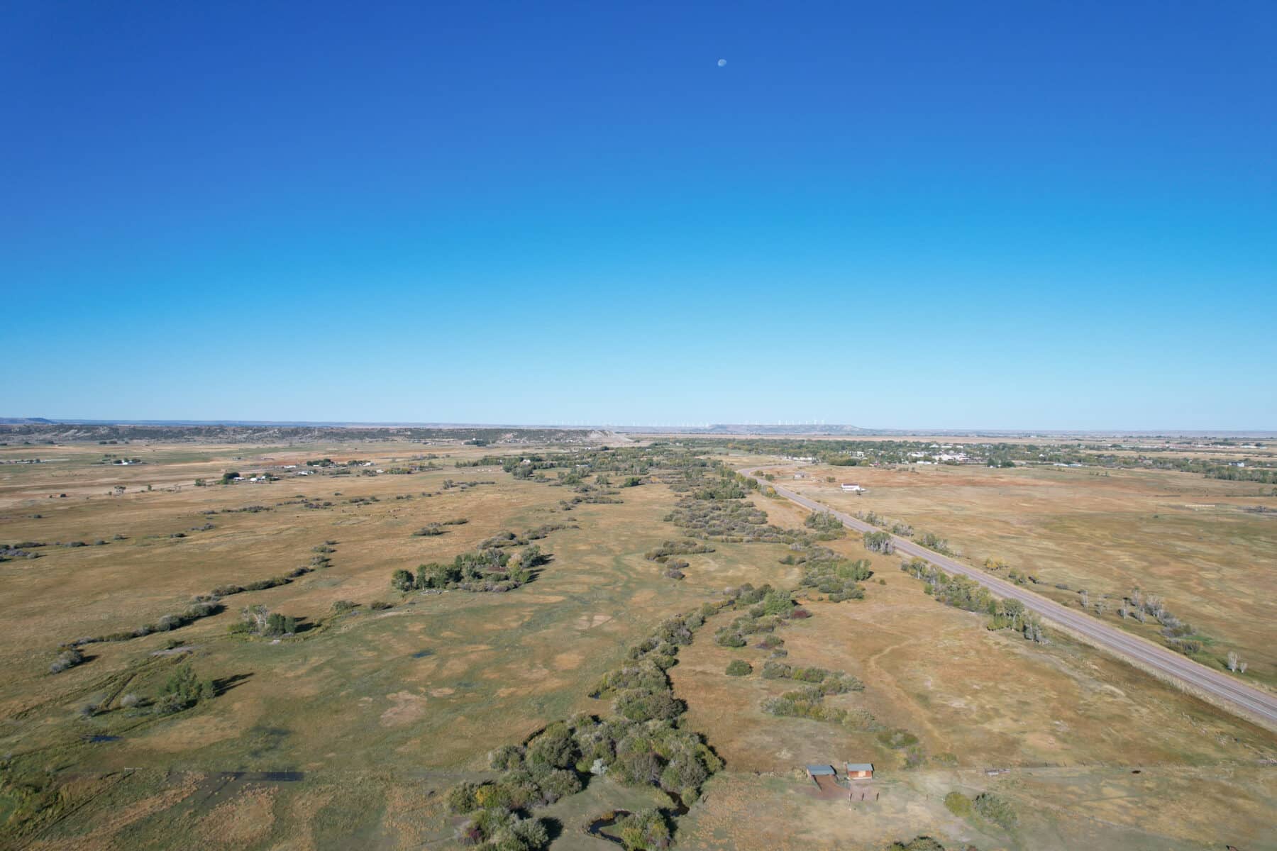 A wide, flat landscape with grassy fields, scattered trees, and a road running through it under a clear blue sky. Ideal as land for sale or hunting property, the distant horizon and faint moon create a serene setting.