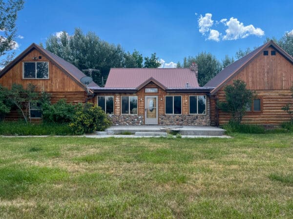 A rustic wooden house with a stone facade, gabled roof, and large windows sits on a grassy lawn under a blue sky—perfect for those seeking a charming ranch for sale surrounded by trees.