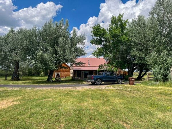 A black pickup truck is parked on a driveway in front of a wooden house with a red roof, surrounded by large green trees. Ideal for recreational land or hunting property, the setting features a blue sky with scattered clouds and a nearby trash bin.