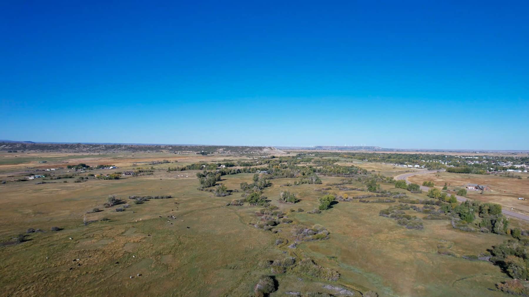 Aerial view of a wide, open rural landscape with green and brown fields, scattered trees, small clusters of houses, and a clear blue sky—ideal for recreational land or as a hunting property.