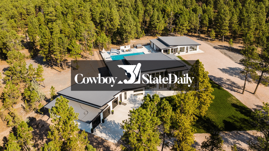 Aerial view of a modern house with a pool, surrounded by trees and scenic land for sale. The Cowboy State Daily logo with a bucking horse icon is overlaid in the center of the image.