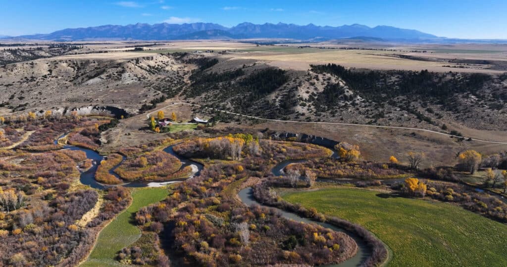 A winding river flows through a scenic valley with patches of green and yellow foliage, surrounded by rolling hills and distant mountains under a clear blue sky—ideal recreational land or hunting property.