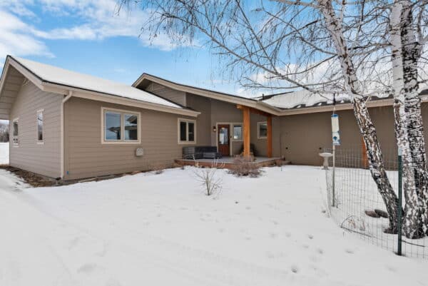 Single-story beige house with a modern design, covered entryway, and large windows, surrounded by snow. A birch tree and bird feeder are visible in the foreground. Ideal for recreational land or as a ranch for sale. The sky is partly cloudy.