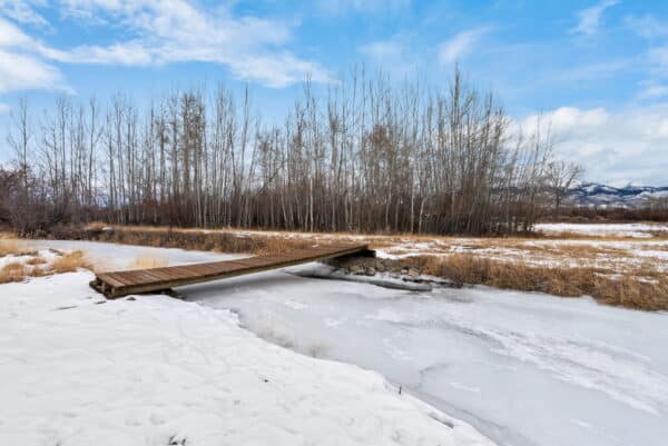 A small wooden footbridge crosses a narrow, frozen stream in a snowy landscape with bare trees and distant mountains—a peaceful scene that could be found on a picturesque cattle ranch or land for sale under blue, cloud-scattered skies.