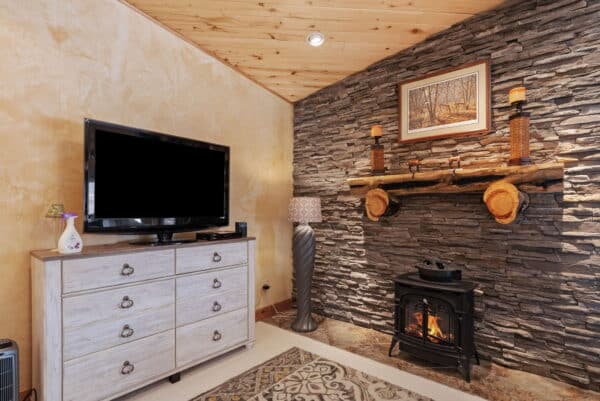 A cozy living room in a hunting property features a TV on a light wood dresser, a modern lamp, a stone accent wall with a rustic mantel, candles, a framed picture, and a small wood stove with a fire burning.