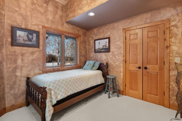 Cozy bedroom with a single wooden bed, patterned bedding, a small gray stool, wood-framed windows, and framed art on textured tan walls. Double wooden doors are closed, and snow is visible outside this inviting cattle ranch retreat.