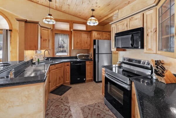 A cozy kitchen with wood cabinets, black countertops, stainless steel appliances, a microwave, oven, toaster, and double sink. Pendant lights hang from a wood-paneled ceiling above a rug on the tile floor—perfect for relaxing after exploring your hunting property.