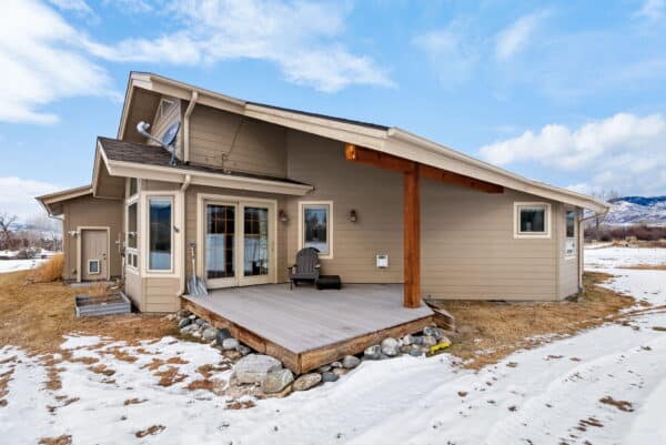 A modern beige house with large windows, a wooden patio, and a slanted roof sits on partially snow-covered recreational land with mountains in the background under a partly cloudy sky.