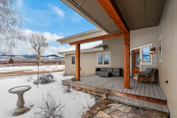 A wooden porch with outdoor seating overlooks a snowy yard and birdbath, with recreational land stretching to mountains and leafless trees in the distance beneath a partly cloudy sky.