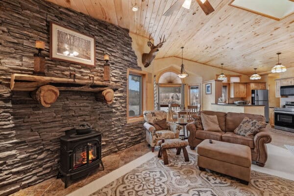 Cozy living room in a hunting property with a stone fireplace, wood-burning stove, deer mount, and rustic wood ceiling. Features a brown couch, armchair, ottoman, and dining area with table near the open kitchen.