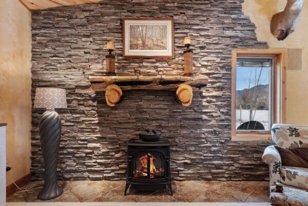 A cozy living room on a cattle ranch features a stone accent wall, wood stove with a fire, rustic wooden mantle, candles, framed artwork, modern floor lamp, snowy window view, and part of a patterned armchair.
