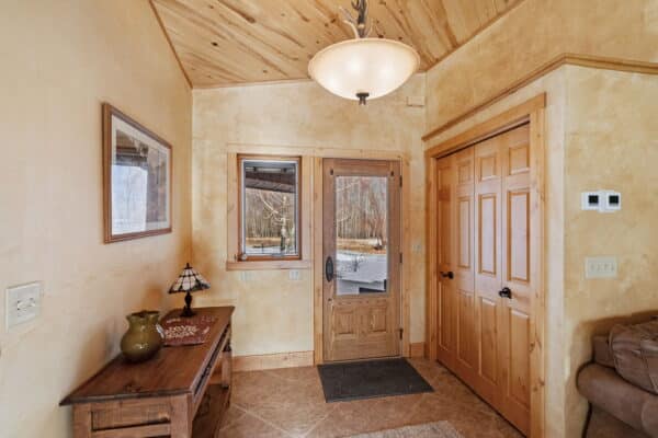 A cozy entryway with a wood-paneled ceiling, beige walls, wooden door with glass panel, small window, double closet doors, wall art, and a console table with lamp and vase—perfect for welcoming guests to your ranch for sale.