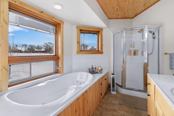 Bright bathroom with a large soaking tub under two windows, a corner shower with glass doors, light wood paneling, and a countertop with drawers—perfect for relaxing after a day on the cattle ranch or exploring nearby land for sale.