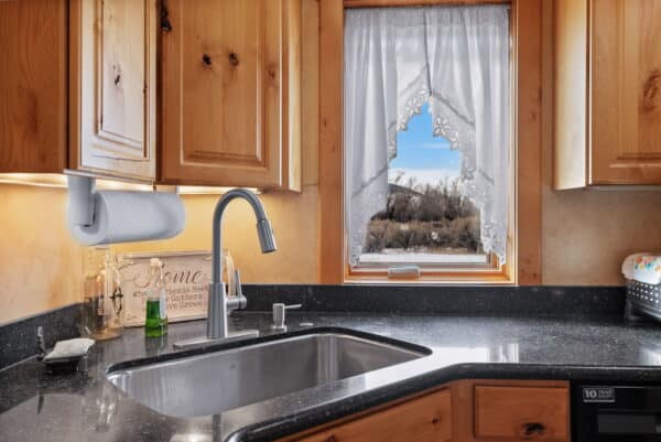 A kitchen sink with a modern faucet sits beneath a window with lace curtains, overlooking a snowy landscape—perfect for a ranch for sale. Wooden cabinets, paper towel holder, soap, and a breadbox rest on the dark countertop.