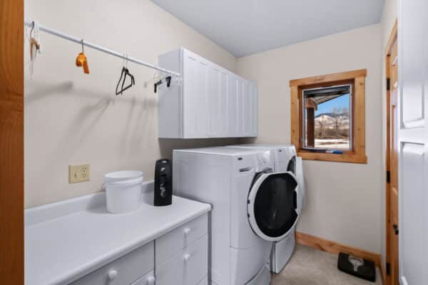 A bright laundry room with white cabinets, washer and dryer, a counter with a container, hanging rods with hangers, and a window with a wooden frame overlooking scenic recreational land.