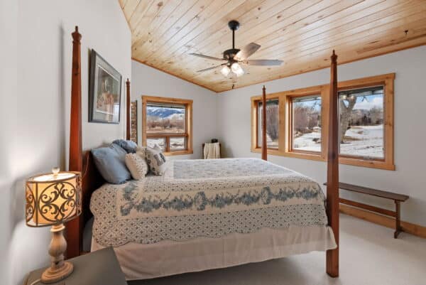 A cozy bedroom with a wooden ceiling, large windows showing a snowy landscape, a four-poster bed with blue and white bedding, a bench, and a lamp on a nightstand. Perfect retreat after exploring nearby hunting property or recreational land.