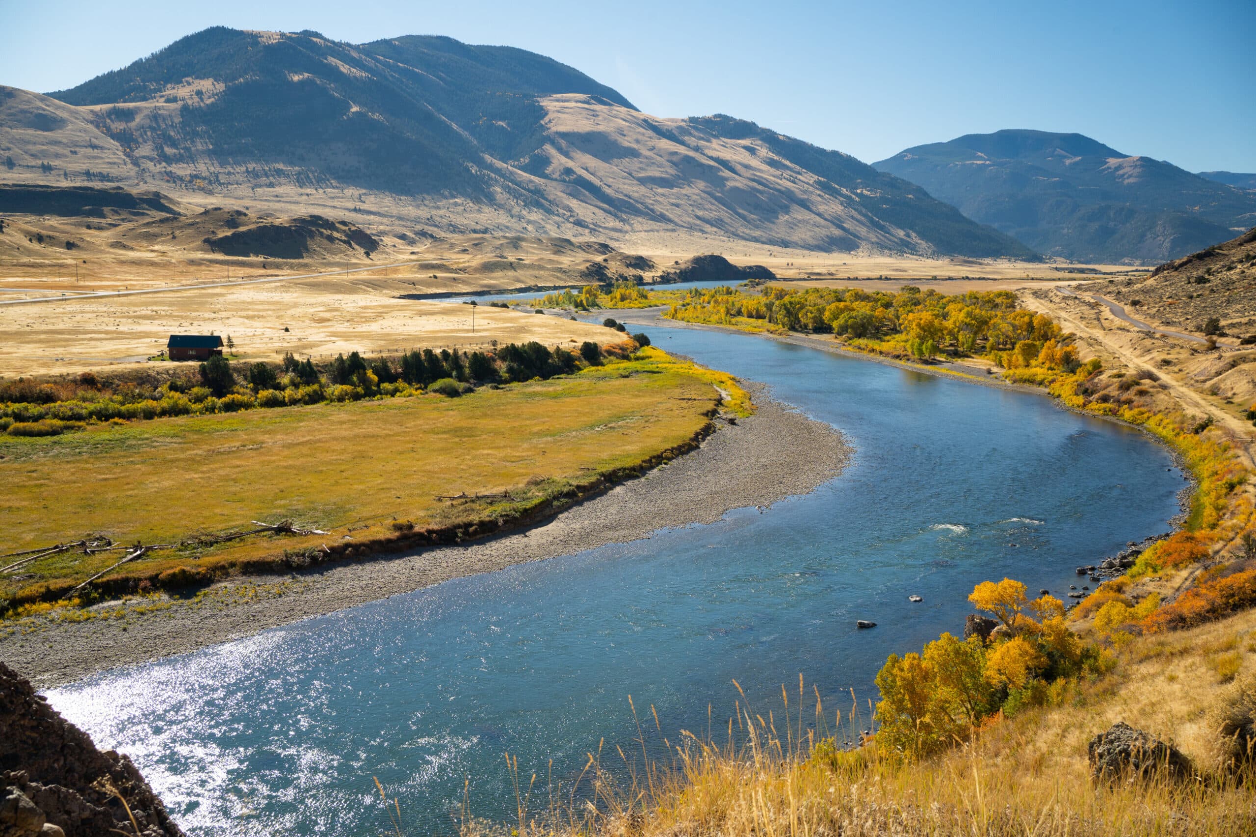 A wide river curves through a sunlit valley with grassy fields and scattered autumn trees, surrounded by rolling hills and distant mountains under a clear blue sky—ideal recreational land or potential cattle ranch.