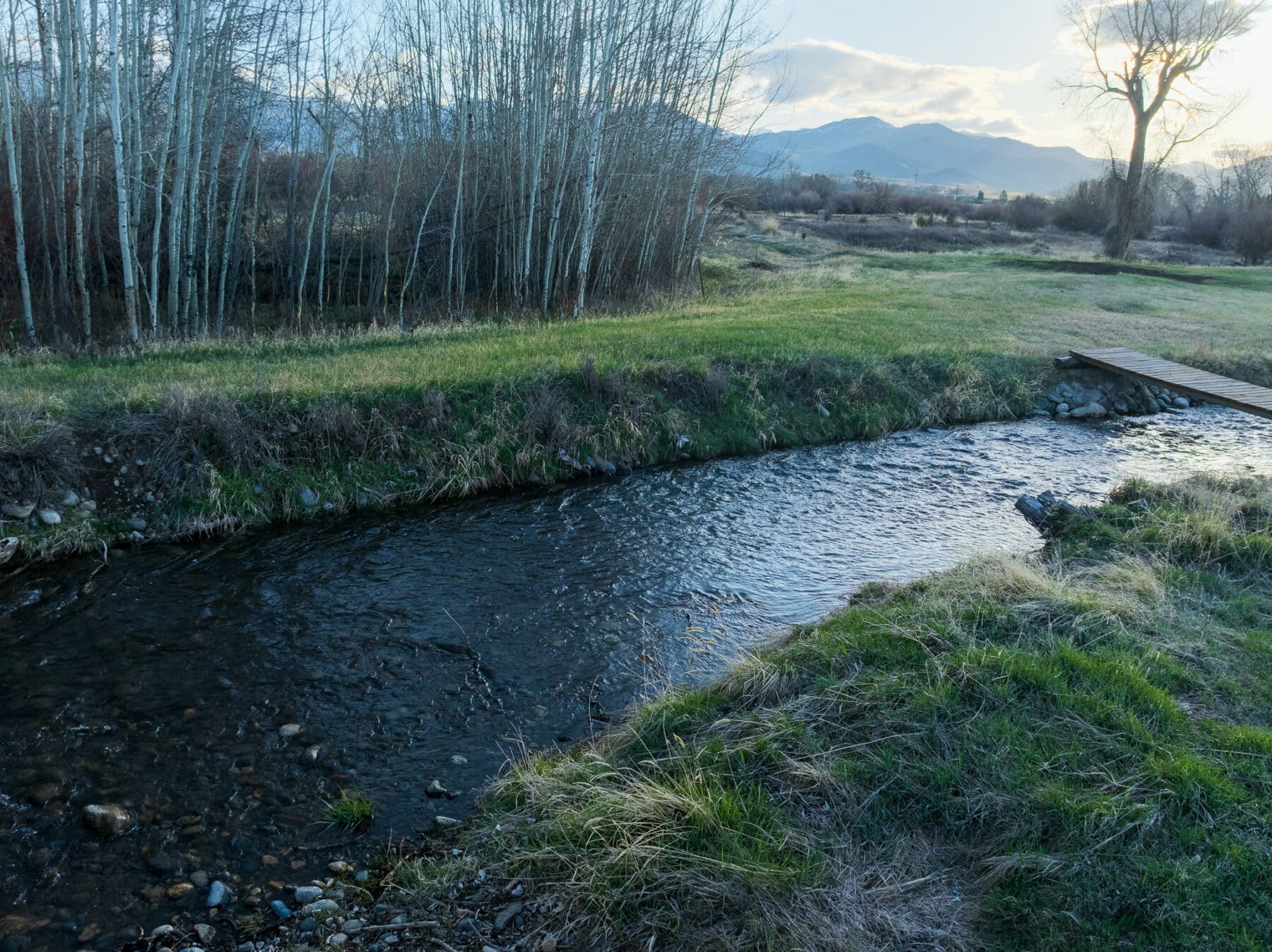 A small creek winds through grassy recreational land with leafless trees on one side and a wooden footbridge. Mountains and a cloudy sky are visible in the background at sunset, highlighting this potential hunting property.