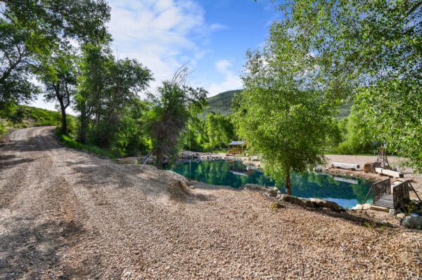 A gravel road curves beside a clear, blue pond surrounded by lush green trees and hills under a partly cloudy sky. A small wooden dock and yellow-roofed structure hint at the charm of this picturesque cattle ranch for sale.