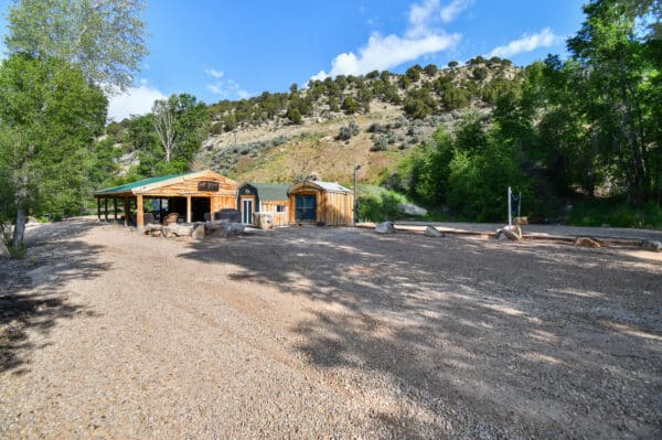 A rustic wooden building with a tin roof sits on a gravel lot surrounded by trees and hills under a blue sky. Perfect as recreational land or hunting property, logs and rocks are placed around the open area in front.