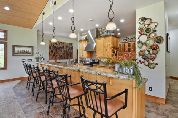 A spacious kitchen with a long granite countertop, five metal barstools, wooden cabinets, stainless steel appliances, and hanging pendant lights. This inviting space is perfect for a cattle ranch or recreational land property. Plants add a touch of greenery.