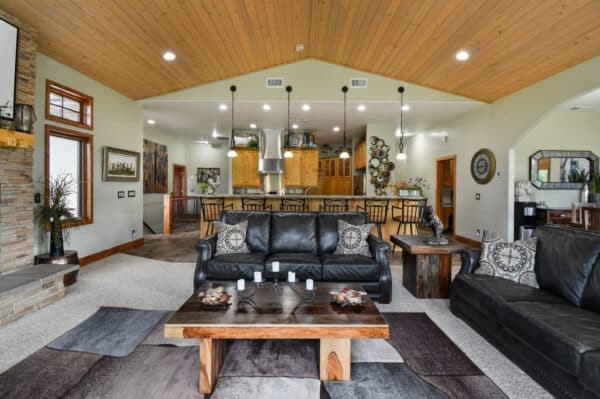 A spacious living room with black leather sofas, a rustic wooden coffee table, and a stone fireplace opens into a modern kitchen with wooden cabinets—ideal for unwinding after exploring your cattle ranch or recreational land.