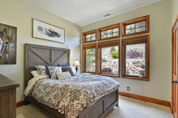 A cozy bedroom with a wooden bed frame, patterned bedding, and a bedside lamp. Large windows with wooden trim overlook a landscaped garden, evoking the charm of a cattle ranch. Artwork decorates the light-colored walls.