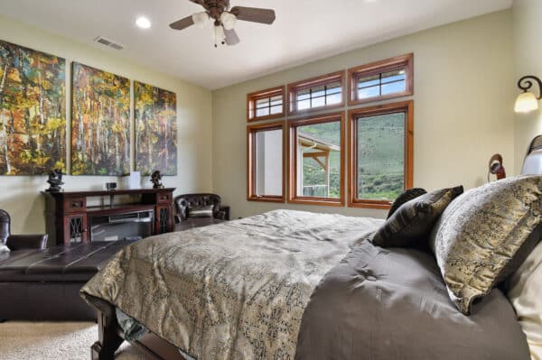 A cozy bedroom with a large bed, patterned bedding, dark wood furniture, leather chairs, and big windows reveals a scenic view of green hills—perfect for anyone searching for a ranch for sale or hunting property.