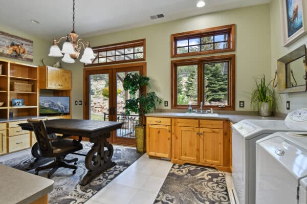 A bright laundry room on a cattle ranch with wooden cabinets, a sink under double windows, a dark wooden table with chairs, a chandelier, washer and dryer, potted plants, and glass doors opening to an outdoor view.