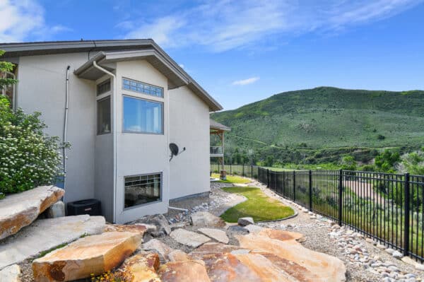 Modern house with large windows and stone landscaping sits on recreational land, overlooking green hills under a blue sky with scattered clouds. A black metal fence runs along the edge of the yard.