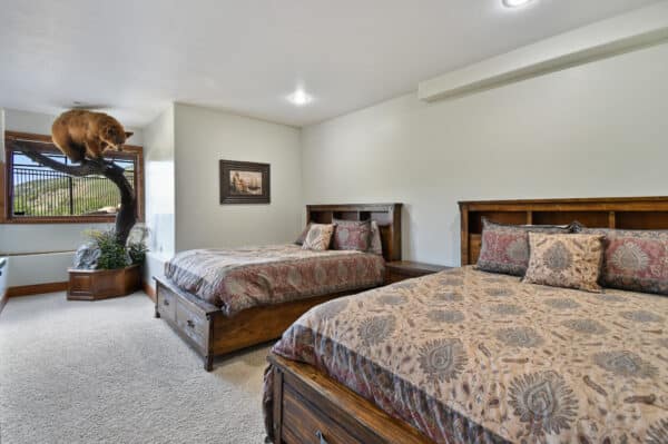 A bedroom on a hunting property with two wooden beds covered in matching patterned bedding, a beige carpet, a framed picture on the wall, and a large mounted bear displayed on a tree near the window.