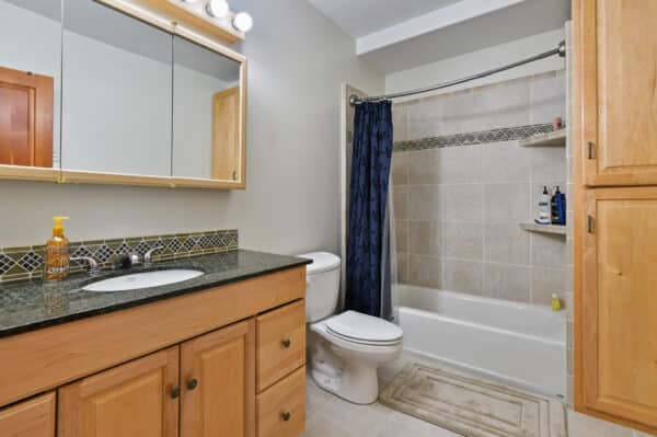 Bathroom with light wood cabinets, black countertop and sink, toilet, bathtub with beige tiles and a dark blue shower curtain, mirrored medicine cabinet, and soap dispenser—perfect for your hunting property or recreational land retreat.