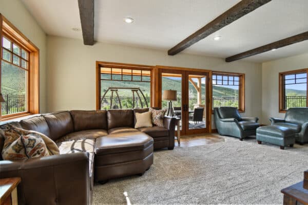 Spacious living room with a brown leather sectional sofa, blue armchairs, large windows, exposed wood beams, and stunning views of green hills—perfect for enjoying the tranquility of your hunting property or recreational land through glass doors to the outdoor patio.