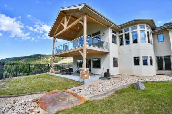 A modern two-story house with large windows, covered balcony with glass railing, and patio on recreational land. The backyard offers a lawn, stone landscaping, and mountain views under a blue sky.