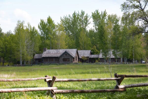 A rustic wooden house with a stone chimney sits in a grassy field, surrounded by tall green trees and partially framed by a split-rail fence—perfect for those seeking recreational land or looking for land for sale.