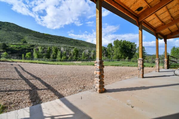 A covered patio with wooden beams overlooks a gravel area, green trees, and a grassy hill under a bright blue sky—perfect for enjoying this scenic recreational land for sale.
