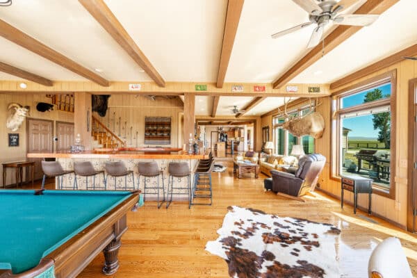 Spacious rustic game room with wood floors, exposed ceiling beams, a pool table, bar area with stools, leather seating, large windows, and cowhide rugs. Perfect for relaxing after a day on your cattle ranch or exploring nearby land for sale. Sunlight streams in from outside.