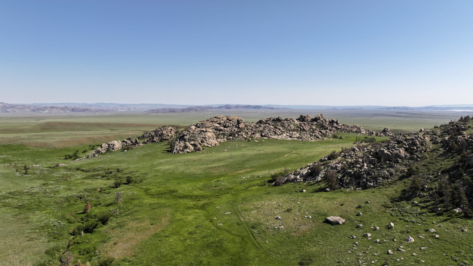 Aerial view of a rocky outcrop surrounded by vast green grasslands under a clear blue sky, with distant hills on the horizon—perfect recreational land or hunting property available for sale.