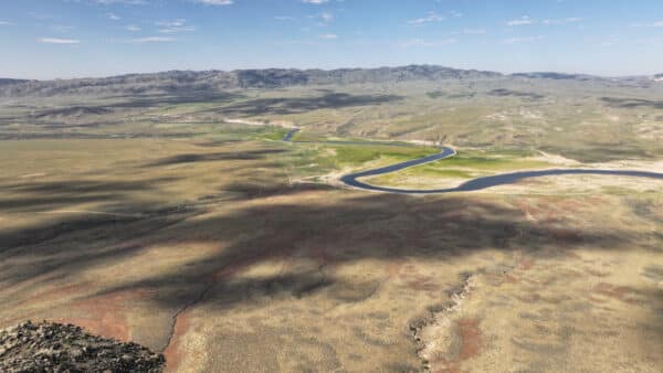 Aerial view of a winding river cutting through a vast, arid plain with scattered greenery, mountains in the background, and blue sky with scattered clouds—ideal recreational land or cattle ranch land for sale.