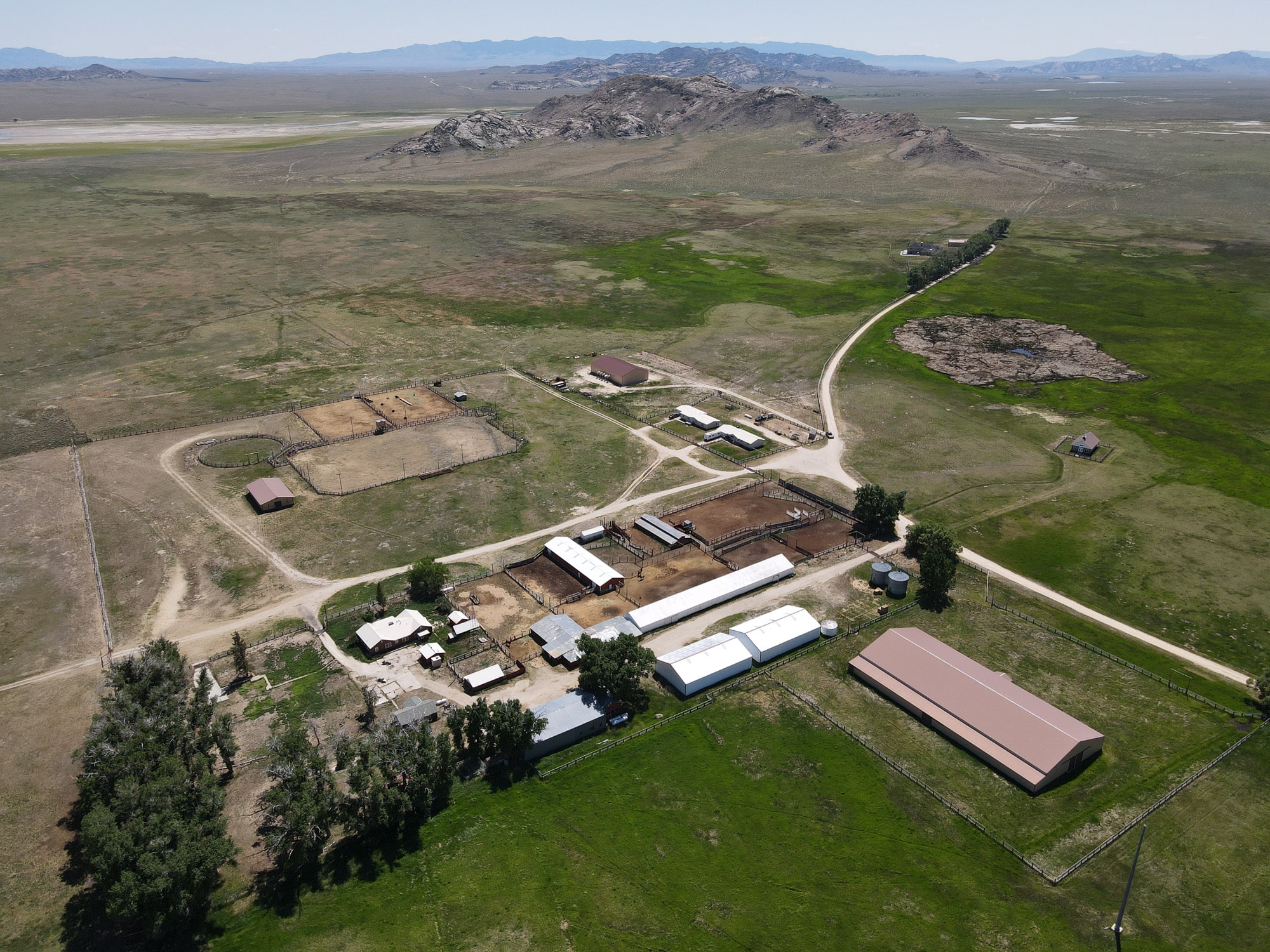 Aerial view of a ranch for sale with several buildings, barns, and fenced areas surrounded by green fields, dirt roads, and distant hills under a clear sky—ideal as hunting property or recreational land.