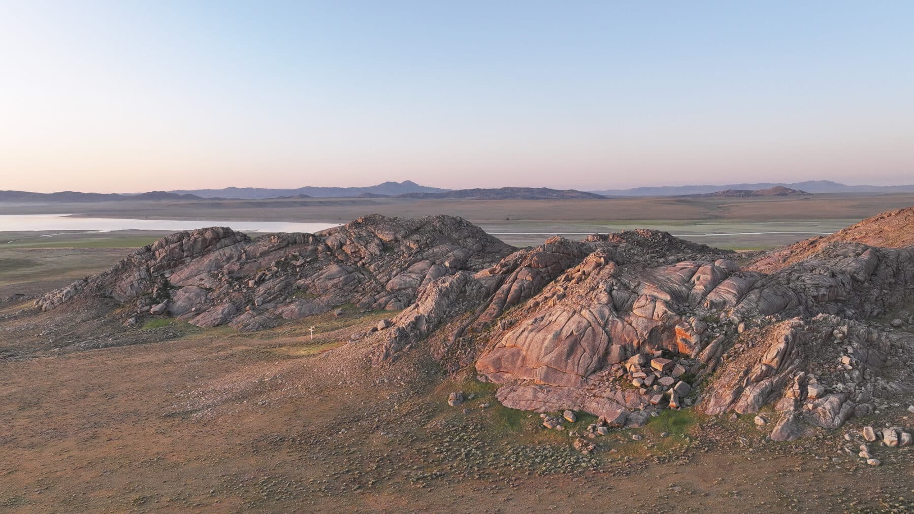 Rocky hills rise from a vast, flat plain under a clear sky at sunset. Soft pink and orange hues highlight the textured rocks of this recreational land, with distant mountains and a body of water visible on the horizon.