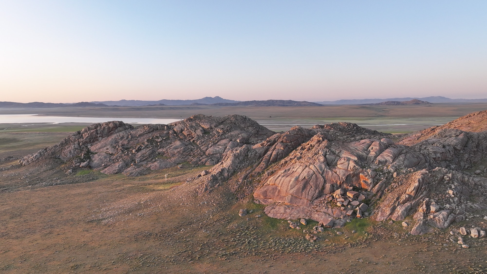 Rocky hills rise from a vast, flat plain under a clear sky at sunset. Soft pink and orange hues highlight the textured rocks of this recreational land, with distant mountains and a body of water visible on the horizon.