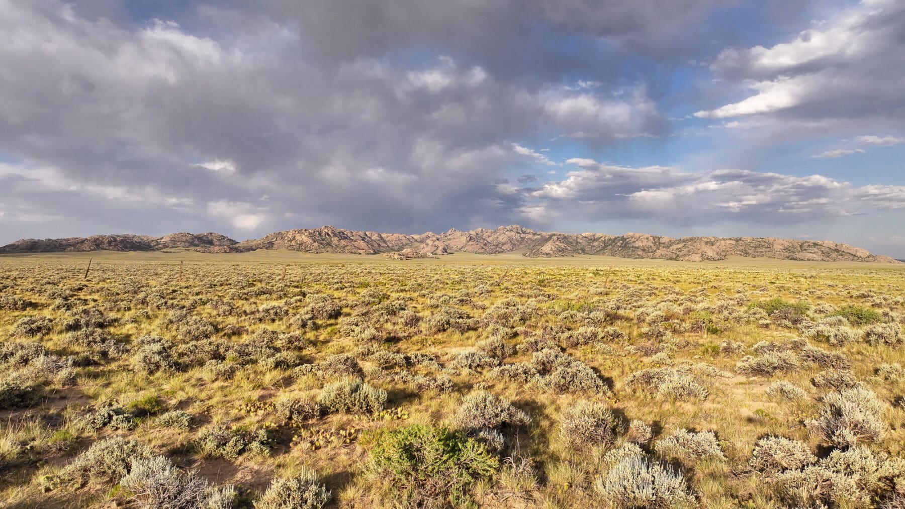 Wide view of open, grassy recreational land with scattered shrubs, leading to distant rocky hills under a partly cloudy sky with sunlight breaking through—ideal hunting property or land for sale.