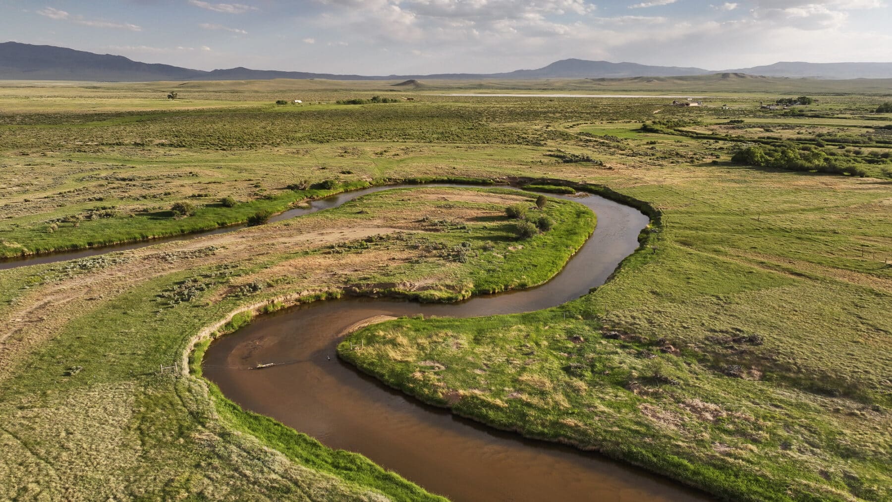 A winding river curves through a green, grassy plain with scattered shrubs. Mountains and a few small buildings are seen in the distance beneath a partly cloudy sky—ideal land for sale or potential hunting property.