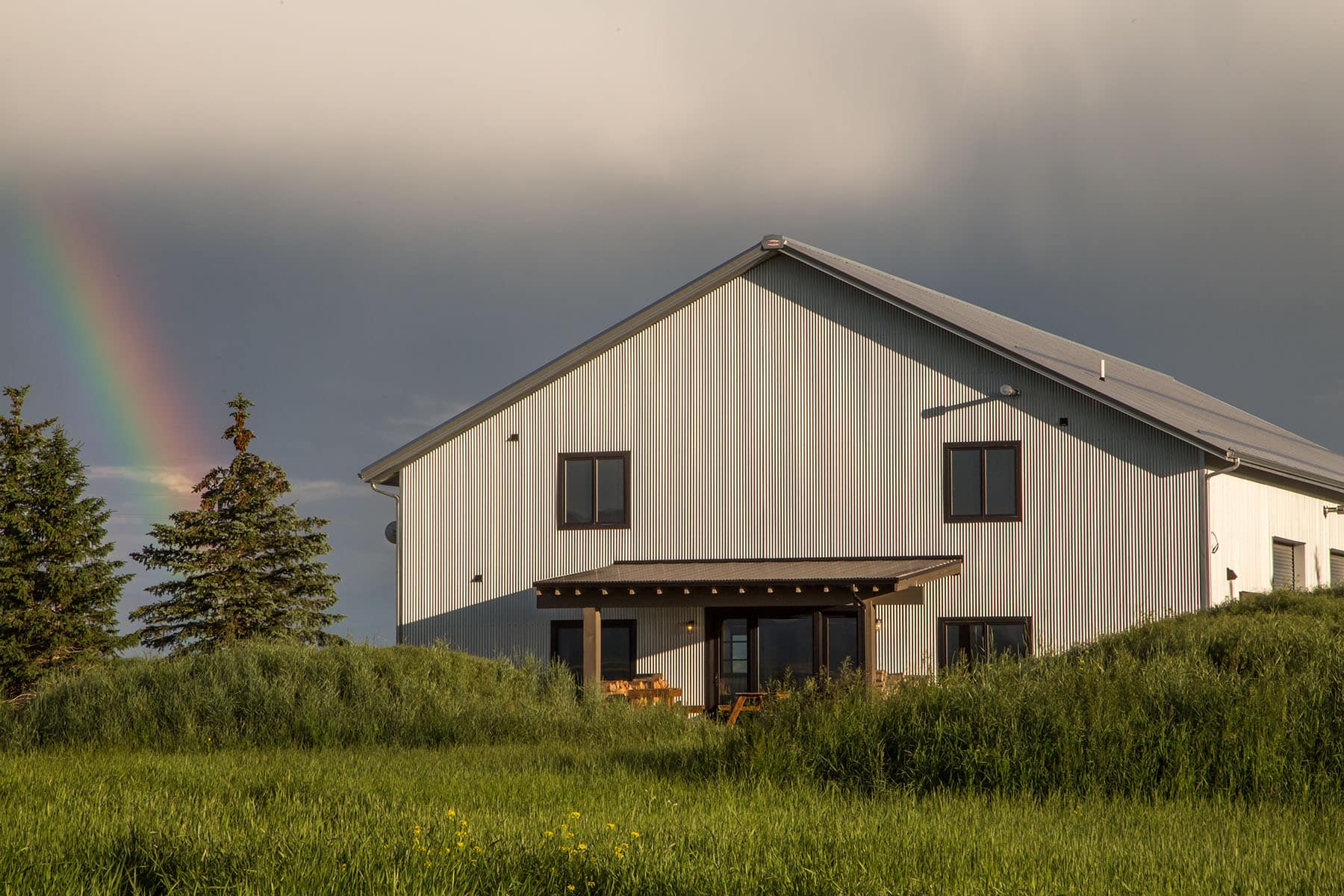 A modern light-gray house sits in a field of tall green grass under a cloudy sky, with a faint rainbow near evergreen trees—an ideal hunting property or ranch for sale.