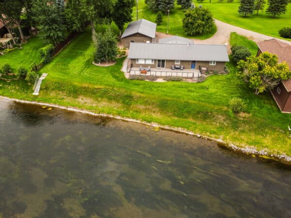Aerial view of a single-story house with a metal roof, surrounded by green lawn and trees next to a calm river. This ranch for sale features a wooden dock extending to the water, with other houses visible nearby.