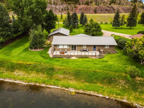 A single-story house with a large deck sits on a well-manicured lawn beside a calm river, surrounded by trees and hills, with a small outbuilding nearby—ideal as hunting property or land for sale.