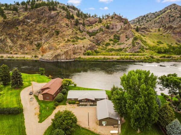 Aerial view of a ranch for sale with manicured lawns beside a river, surrounded by rocky hills and trees under a partly cloudy sky—ideal recreational land in a scenic setting.