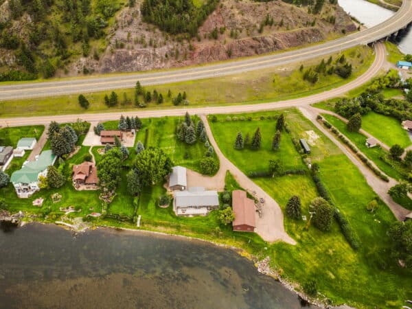 Aerial view of houses with large green lawns beside a river, bordered by trees, a road, and a highway along a hillside—ideal as hunting property or perfect ranch for sale in a serene natural setting.